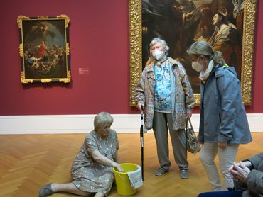 2 Besucherinnen neben der sitzenden "Putzfrau", an der Wand hängen Bilder