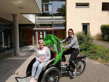 Eine Frau sitzt auf einer Fahrradrikscha. Eine jüngere Frau sitzt auf dem Sattel.