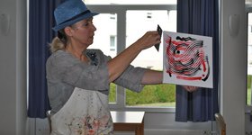 Künstlerin Bjerstedt mit blauem Hut hält Bild in Pouring-Technik hoch