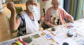 2 ältere Frauen bemalten Steine und lachen in die Kamera